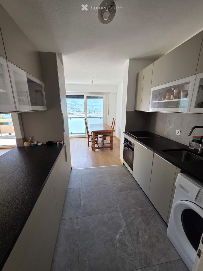 Modern hallway leading to a kitchen and dining area, with grey tiled floor, white cabinets, and a view of a balcony.