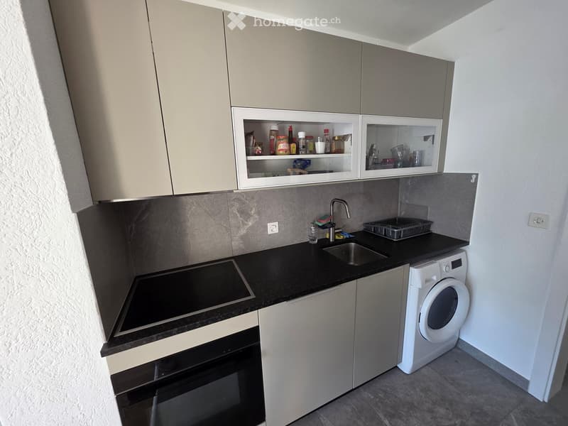 Modern kitchen with light grey cabinets, black countertops, a sink, a washing machine, and an oven.