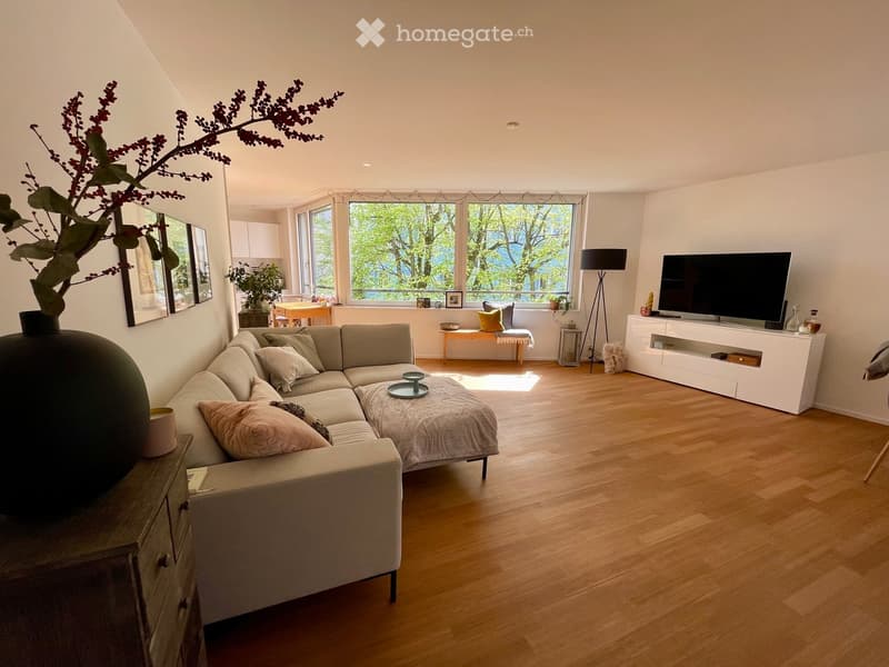 Modern living room with light wooden flooring, large windows, a sectional sofa, coffee table, TV stand, floor lamp, and decorative vases.