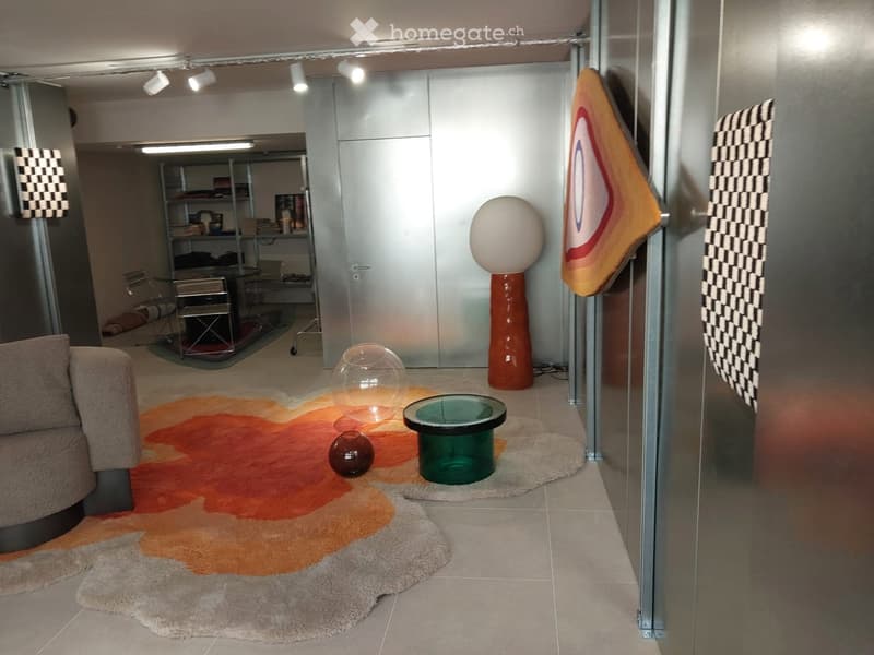 Modern living room with a large orange and beige rug, a grey sofa, a green glass table, decorative vases, and a colorful abstract sculpture. The room features sleek, contemporary furniture and a glass wall separating it from another area.