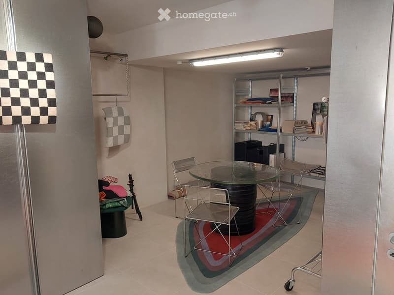 A small, furnished room with a round glass table, four chairs, a shelf unit with folded clothes and books, and a decorative lamp near the entrance.