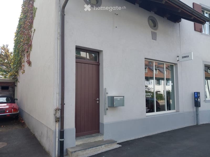 Two-story building with a light-colored facade, a brown front door, a window with a sign reading "DONKKA", and a small parking area with a red car.