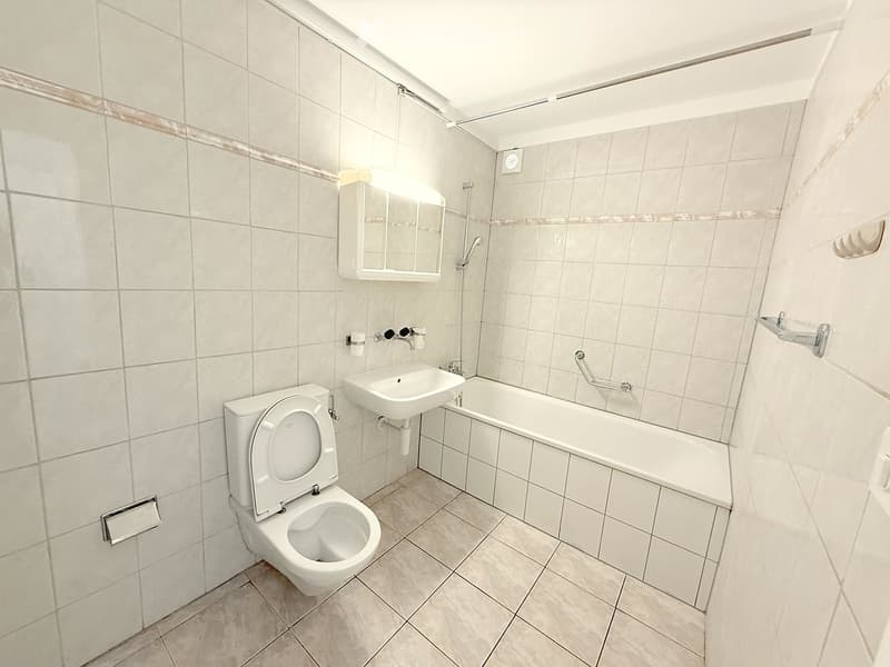 Modern bathroom with white tiled walls and floor, featuring a toilet, sink, bathtub, and a mirror above the sink.