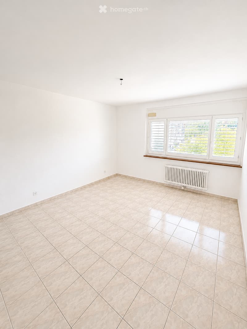 Empty room with white walls, tiled floor, and three windows with blinds. A radiator is visible under the windows.