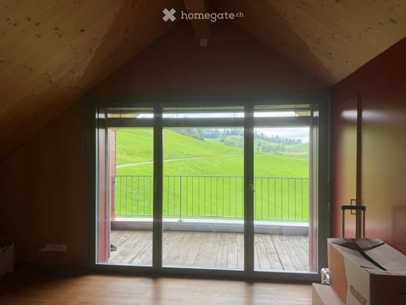 Empty attic room with wooden floors, large sliding glass doors opening to a wooden deck and scenic green landscape view.