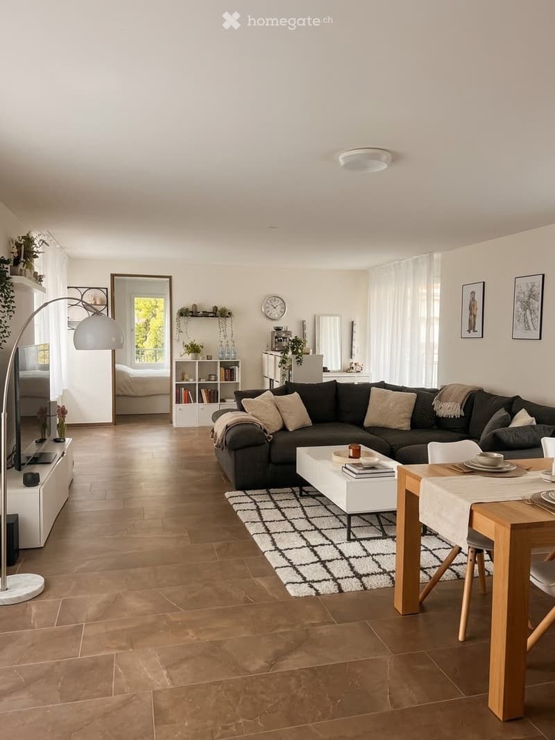 Modern living room with a dark grey sectional sofa, white coffee table, dining table with chairs, bookshelf, floor lamp, and decorative items.