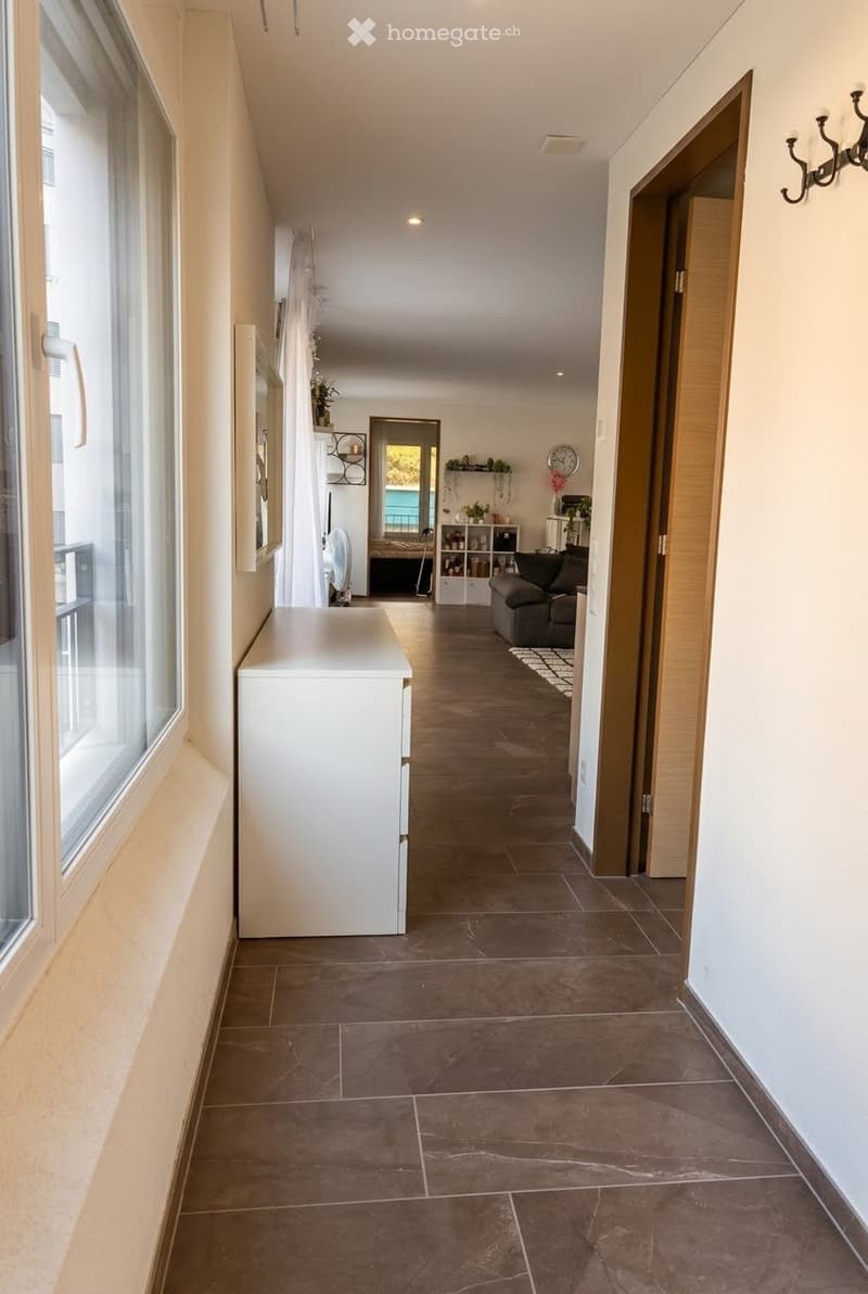A modern hallway with tiled flooring, white walls, and large windows on one side. The hallway leads to a living area with a sofa, coffee table, and decorative plants.