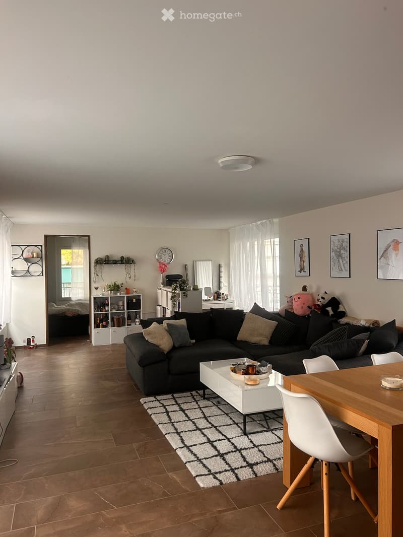 Modern living room with a black sectional sofa, white coffee table, dining table with chairs, and a shelf with decorative items. The room features wooden flooring, a patterned rug, and large windows with white curtains.