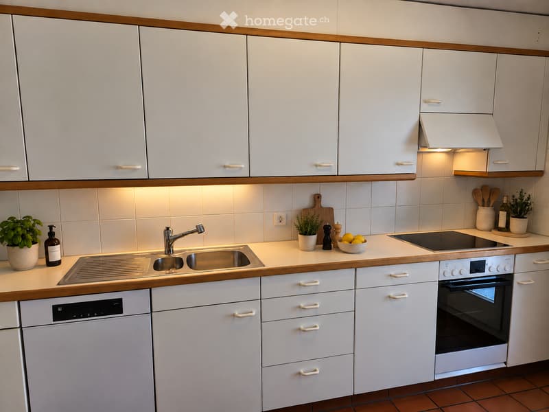 Modern kitchen with white cabinets, stainless steel sink, oven, and dishwasher. Countertop features a bowl of lemons, cutting board, potted plants, and a bottle of olive oil.