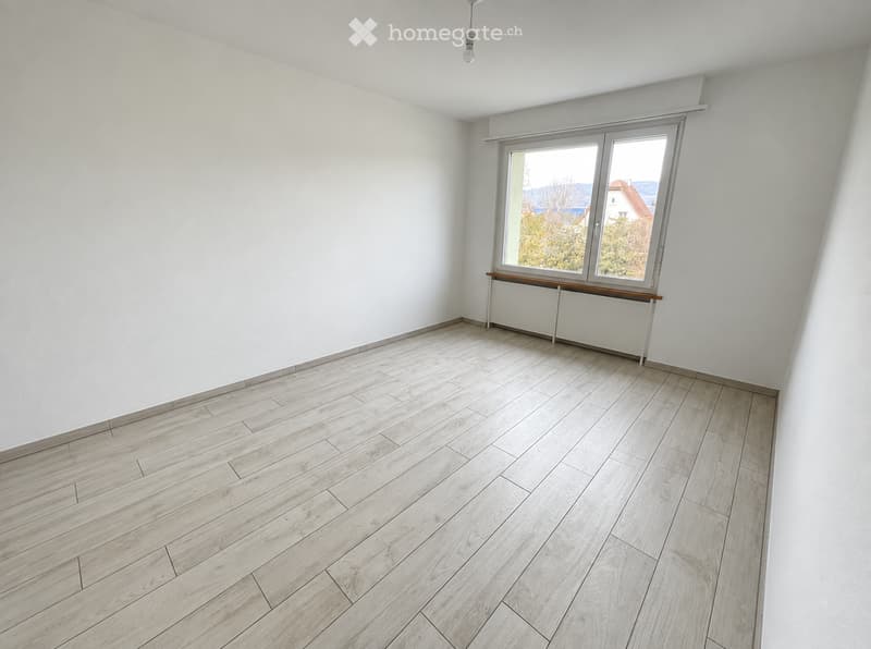 Empty room with light wooden flooring, white walls, and a large window with a simple wooden shelf below it.