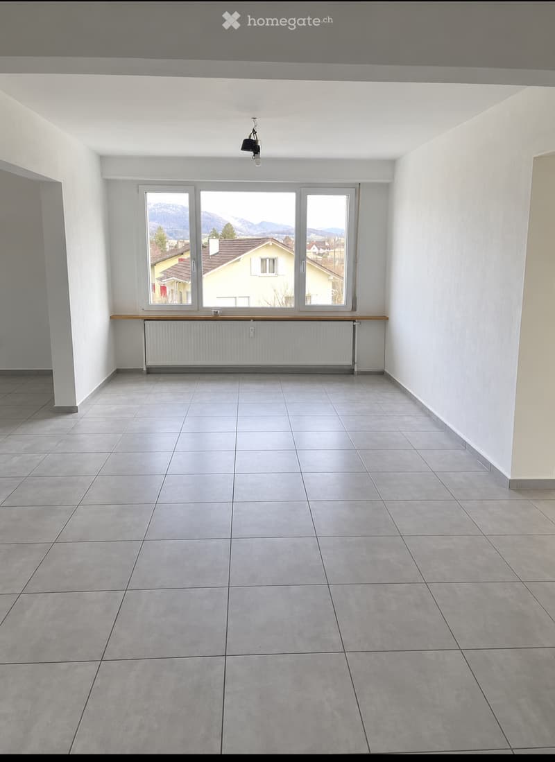 Empty room with tiled floor, white walls, large windows with a view of mountains, radiator, and a small wooden shelf.