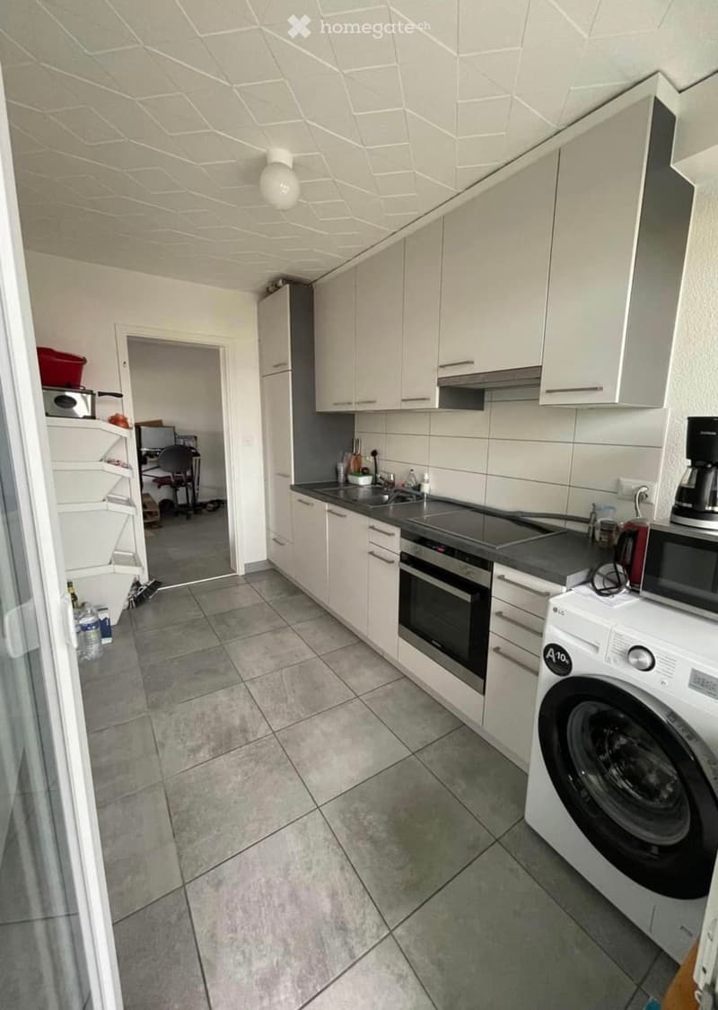 Modern kitchen with white cabinets, stainless steel appliances, and a tiled floor. Includes an oven, washing machine, microwave, and a small refrigerator in the adjacent area.