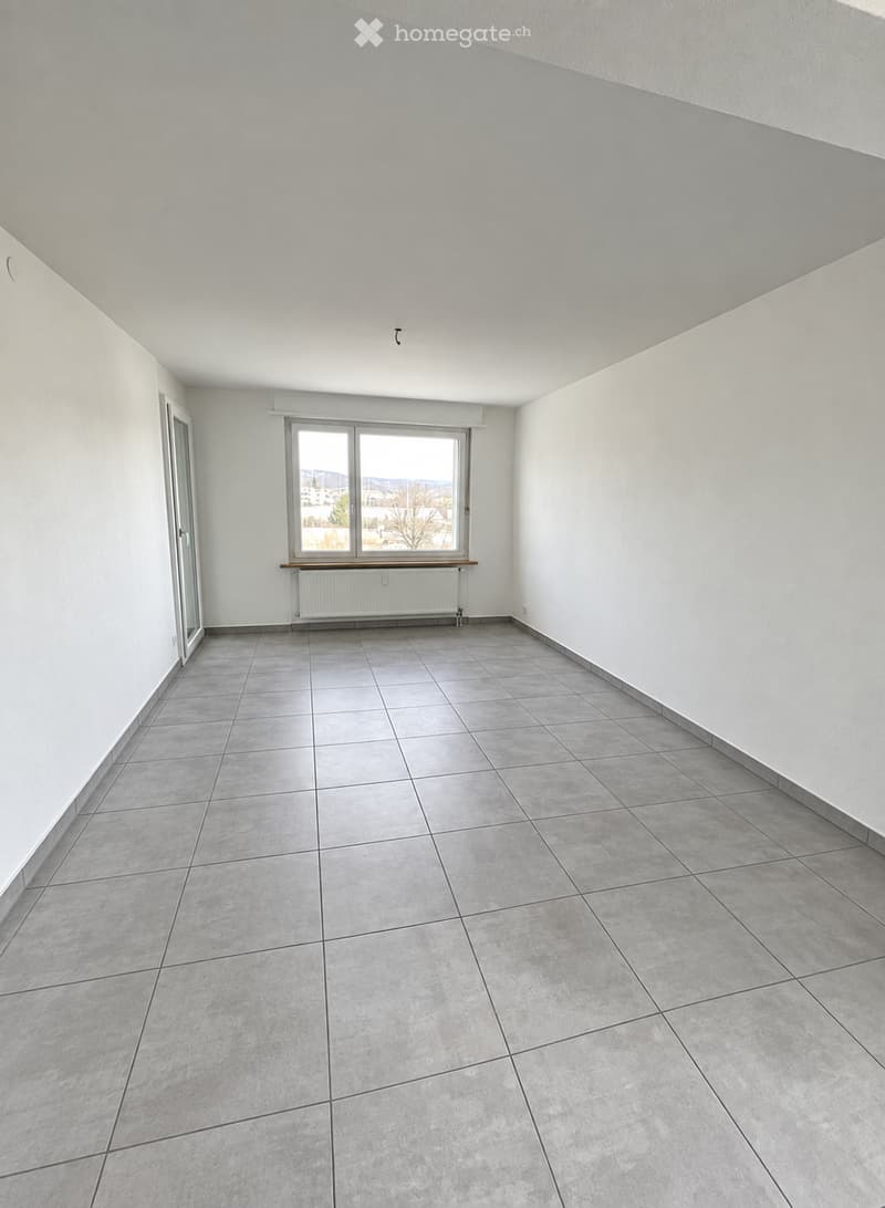 Empty room with tiled floor, white walls, large window with view, radiator, and door.