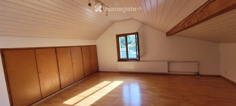 Empty room with wooden flooring, light-colored walls, and a large window letting in natural light. Features built-in wooden cabinets and a radiator under the window.