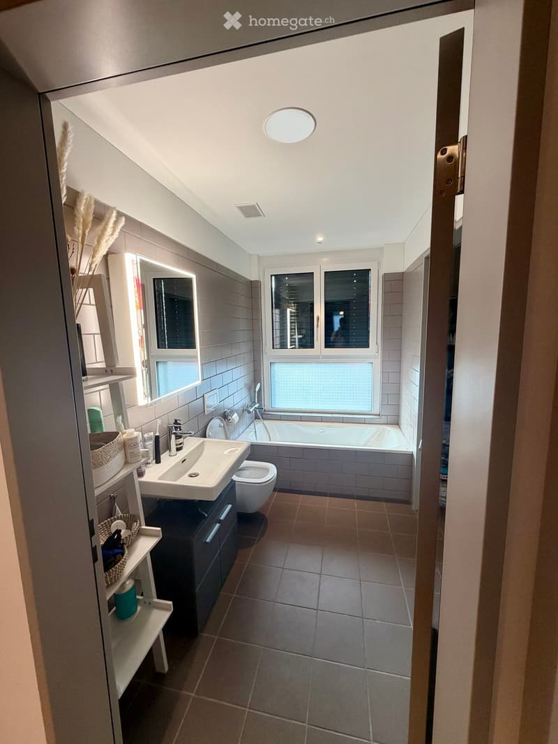 Modern bathroom with a sink, toilet, bathtub, and large window. Includes a mirrored cabinet, shelves with baskets, and various toiletries.