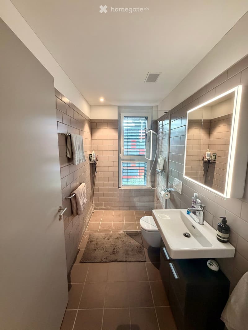 Modern bathroom with beige tiles, a large sink with a modern faucet, a toilet, a shower area with a glass enclosure, a large illuminated mirror, and towels on the wall.