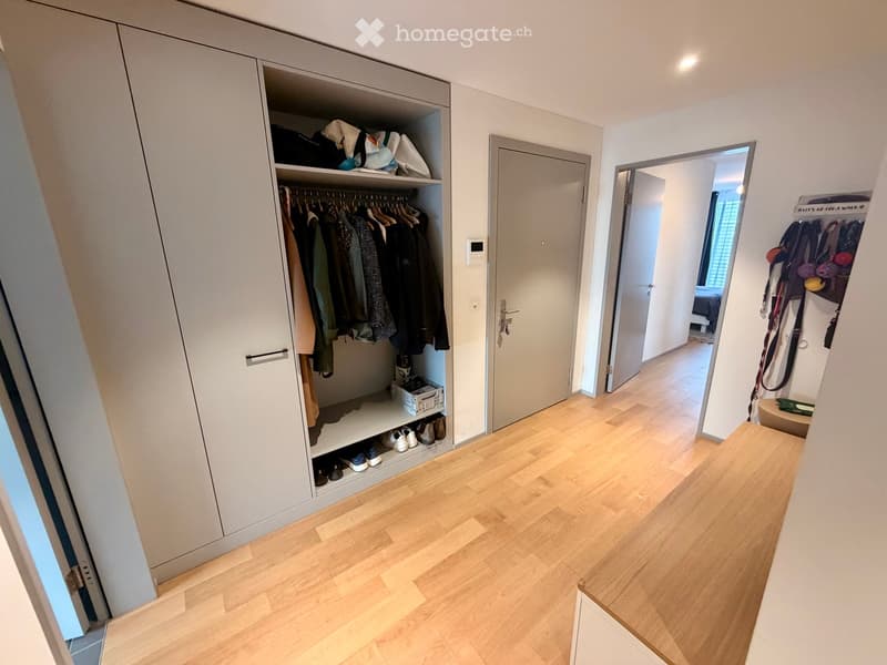 A modern hallway with light wooden flooring, white walls, and built-in closet with hanging clothes and shoes. A door leads to another room, and a small shelf with items is visible in the background.