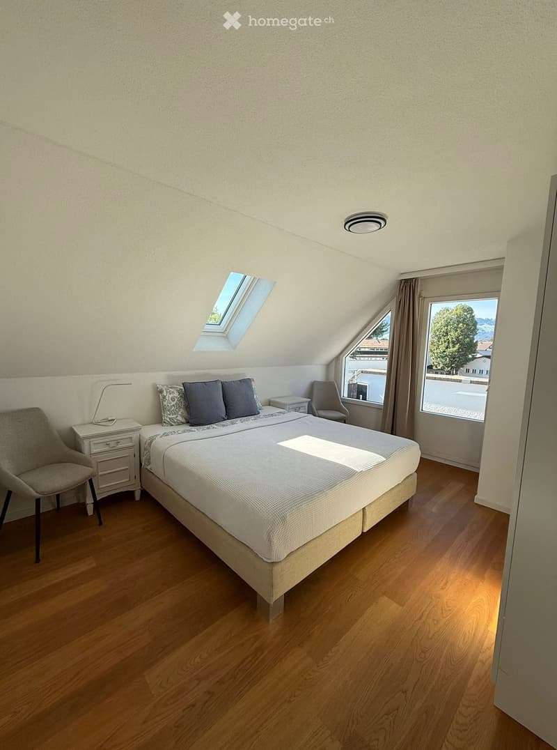 Cozy attic bedroom with wooden flooring, large bed, nightstand, chair, and skylight window offering natural light.