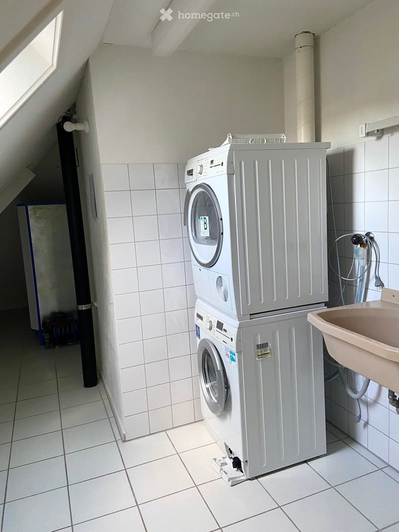 A small bathroom with a washing machine and a sink, featuring white tiled walls and a skylight window.