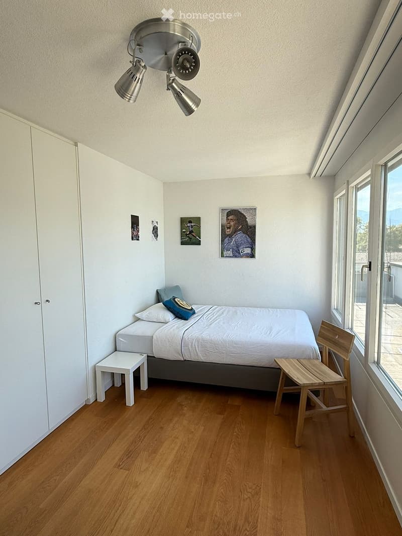 A small, modern bedroom with a single bed, white linens, a wooden floor, a small white stool, a wooden chair, and a small table. The room features large windows with a view and several framed pictures on the wall.