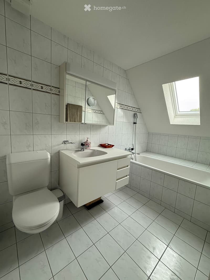 Modern bathroom with white tiles, a toilet, a sink with a cabinet, a bathtub, and a mirror above the sink. The room features a window letting in natural light.