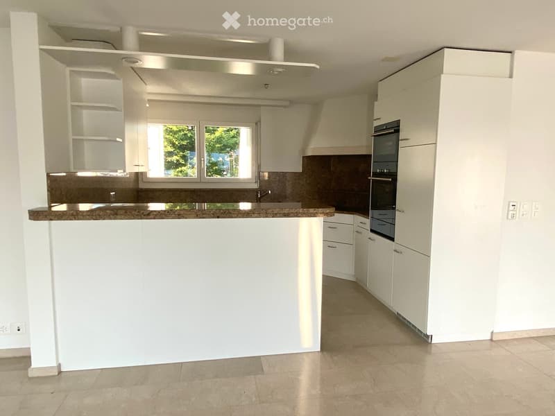 Modern kitchen with white cabinets, granite countertop, and built-in oven. Features a central island and large windows with a view of greenery.