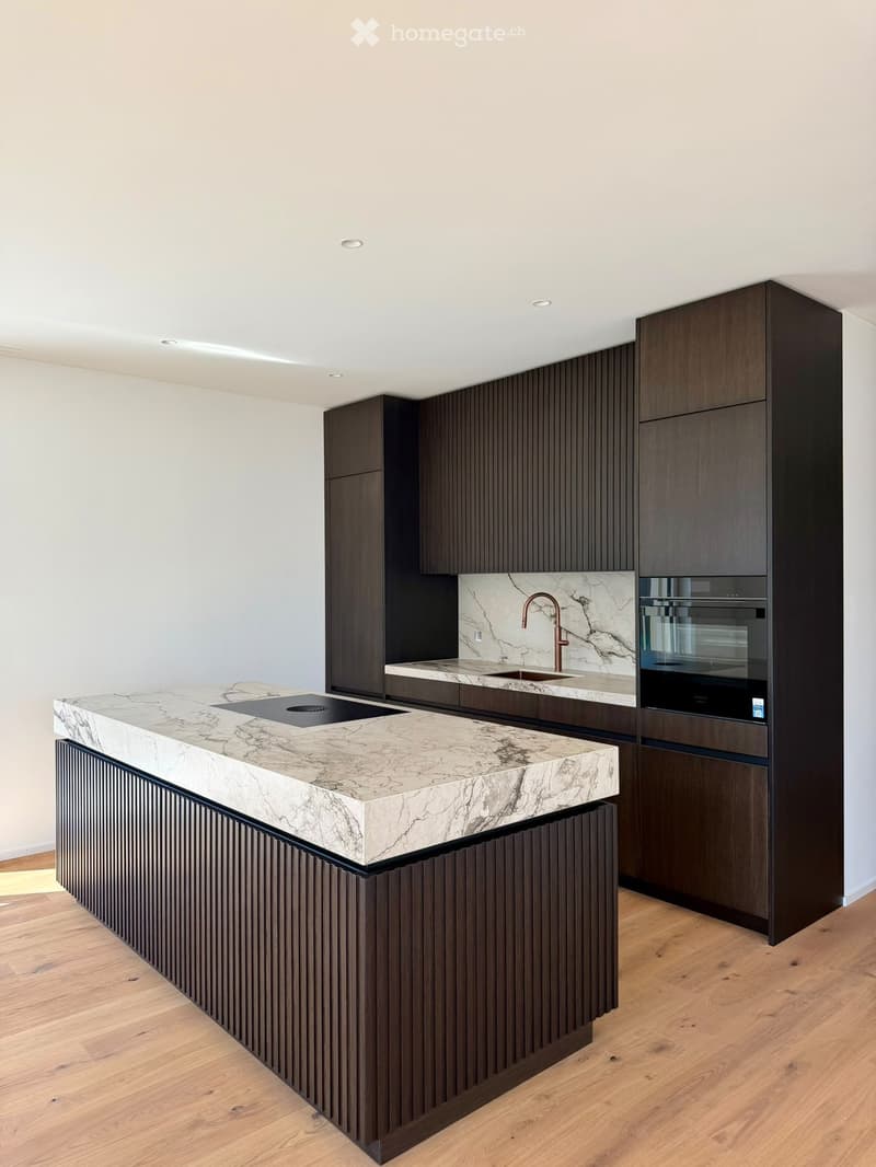 Modern kitchen with a large island, marble countertops, dark wood cabinetry, and built-in appliances including an oven and sink.