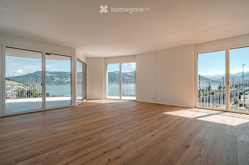 Bright, empty room with wooden flooring and large glass sliding doors leading to a balcony with scenic mountain and lake views.