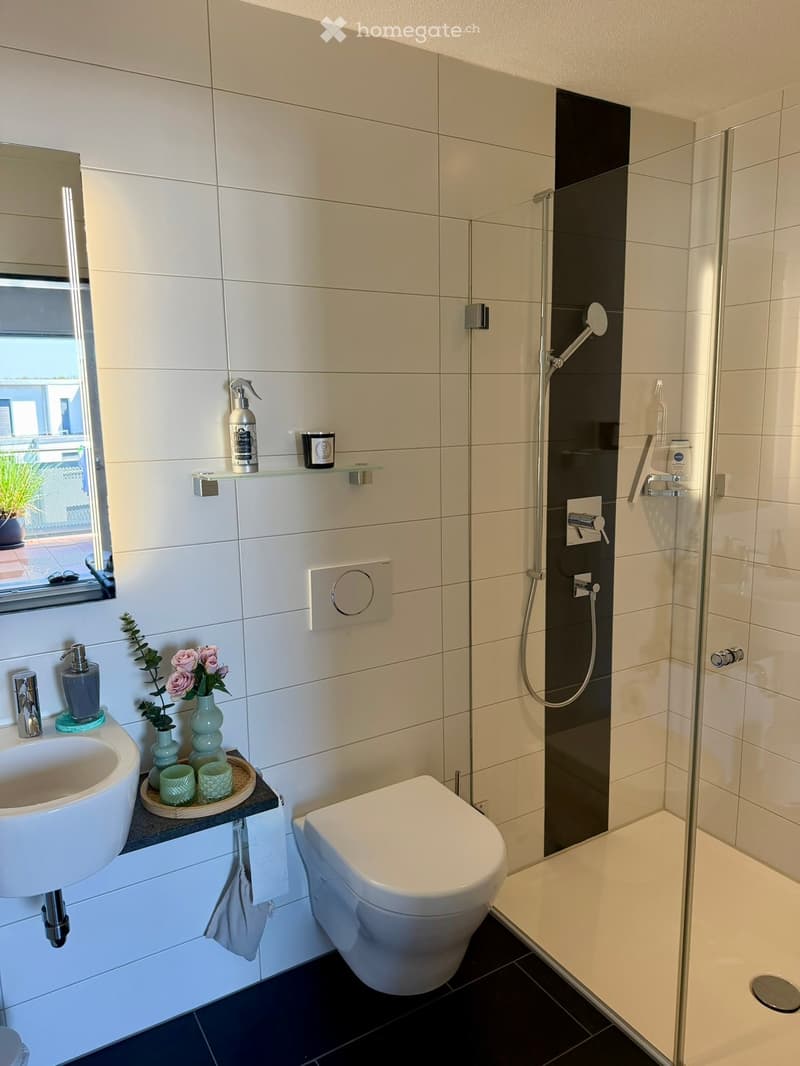 Modern bathroom with white tiles, a glass-enclosed shower, a white toilet, a sink with a modern faucet, and decorative items including a vase with pink flowers, cups, and a towel.