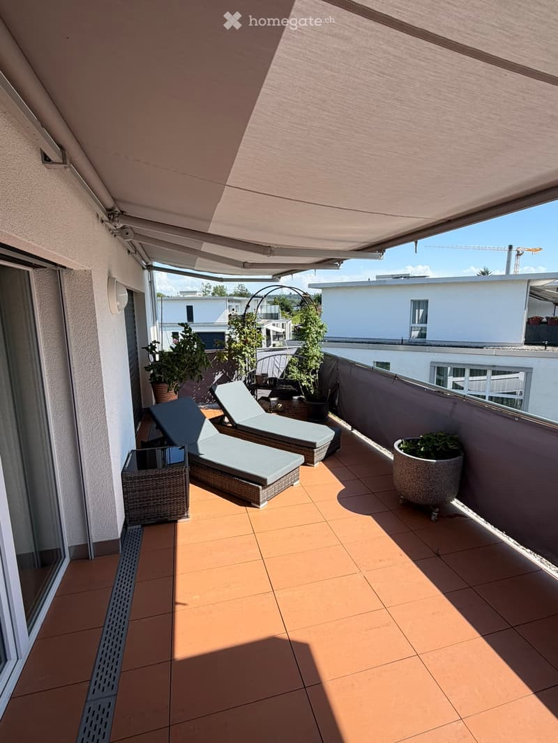 Balcony with sun loungers, a small table, and potted plants, featuring a retractable awning and a view of neighboring buildings.