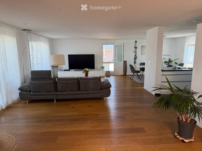 Modern living room with hardwood flooring, large windows, and a balcony. Features a grey sectional sofa, flat-screen TV, floor lamp, table lamp, vase with flowers, and a potted plant.