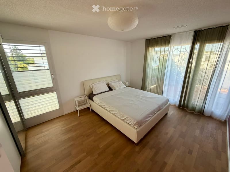 Modern bedroom with a large bed, white bedding, a nightstand, and large windows with blinds and curtains. The room features wooden flooring and a round ceiling light.