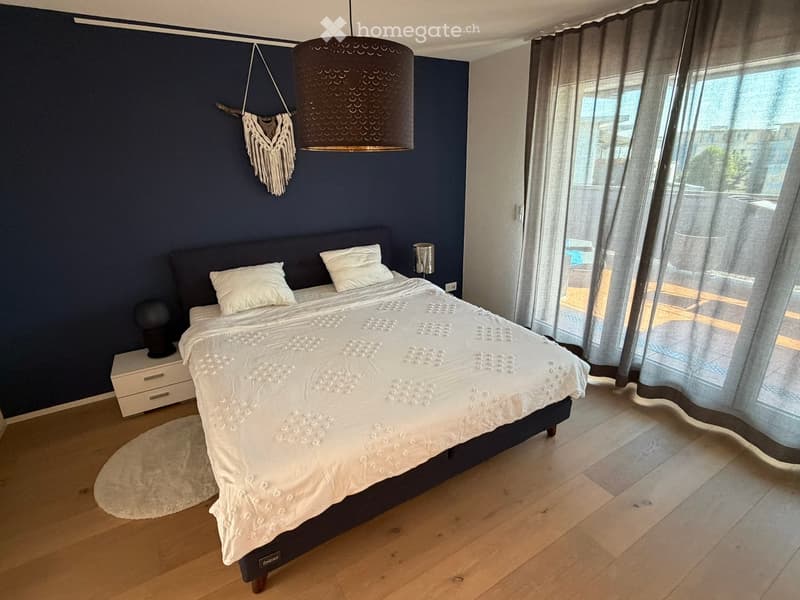 Modern bedroom with a large bed, white bedding, nightstand, and decorative wall hanging. Large windows with sheer curtains let in natural light.