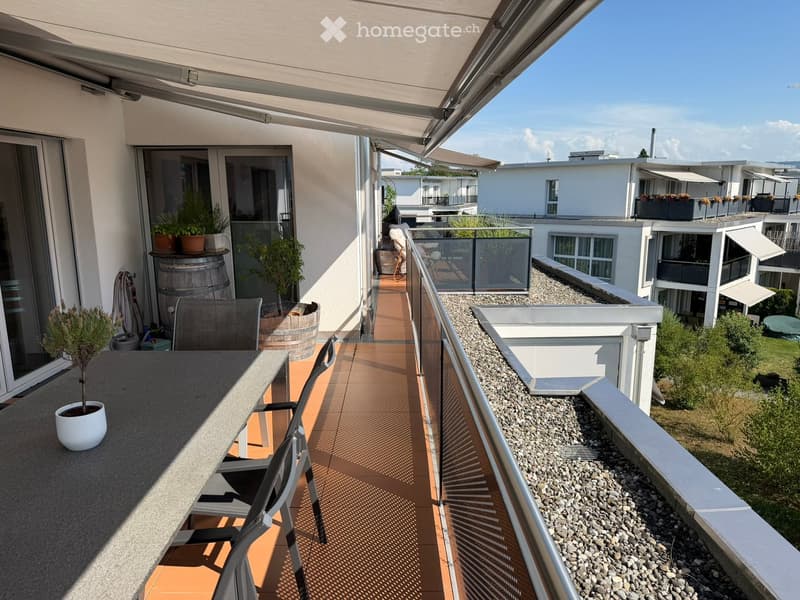 Balcony with a table, chairs, potted plants, and a view of the surrounding buildings and greenery.
