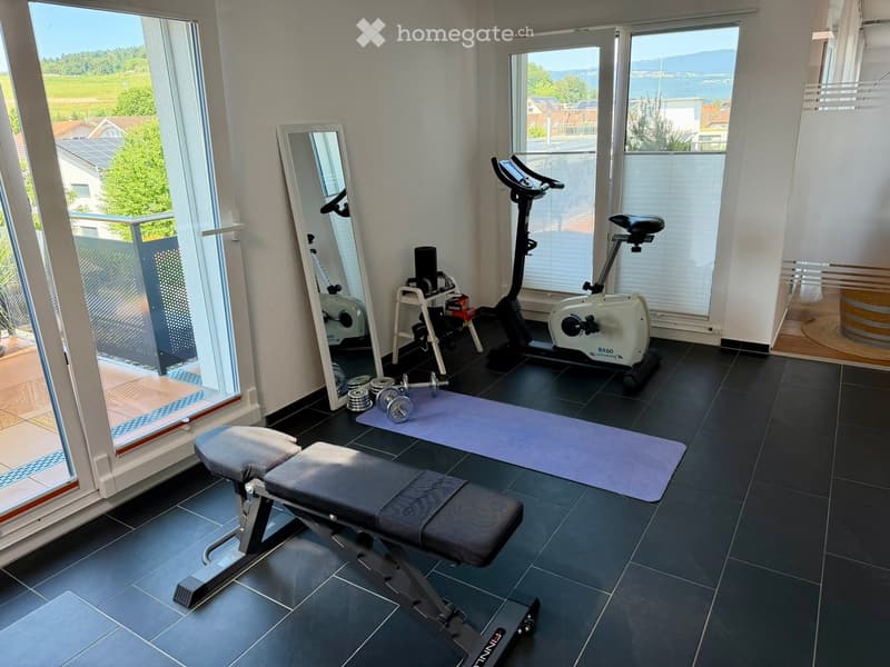 Well-lit fitness room with exercise bike, bench, dumbbells, mirror, and yoga mat. Large windows offer scenic views.