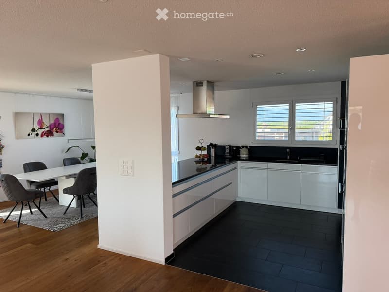 Modern kitchen with white cabinets, black countertops, and a dining area with a white table and grey chairs. The kitchen features a stainless steel hood, sink, and various appliances.