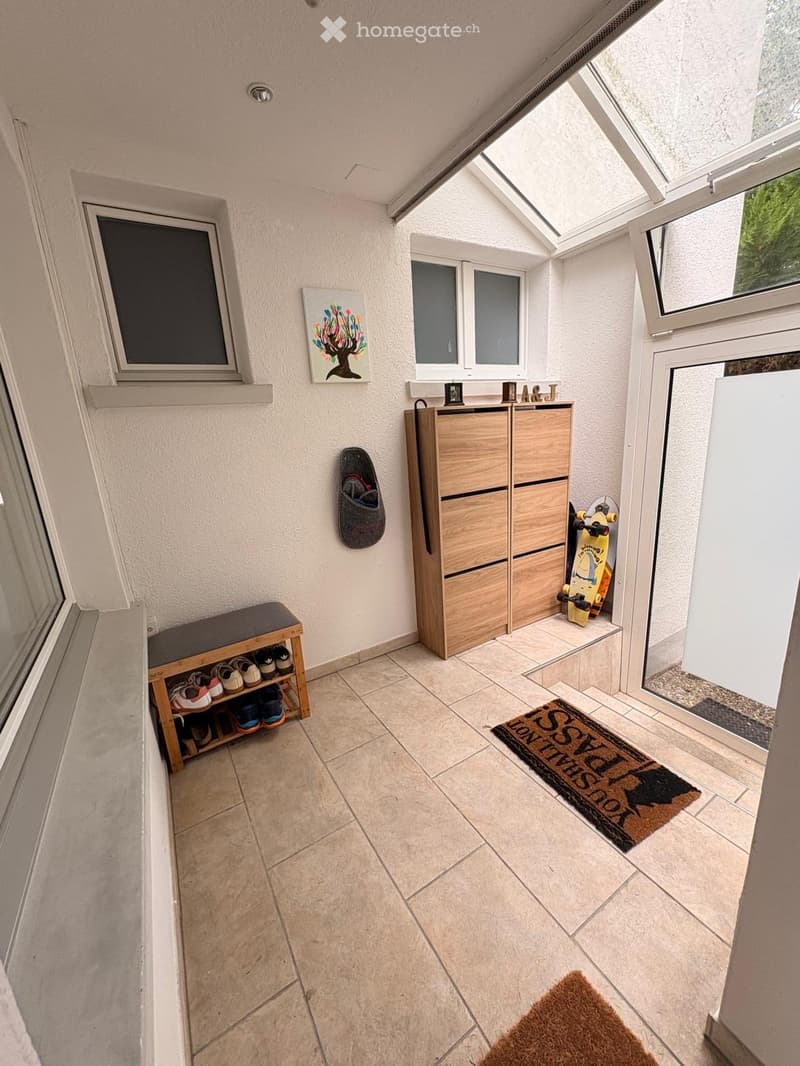 Bright hallway with tiled floor, wooden shoe rack, storage unit, and a small bench. Features a doormat and a painting on the wall.