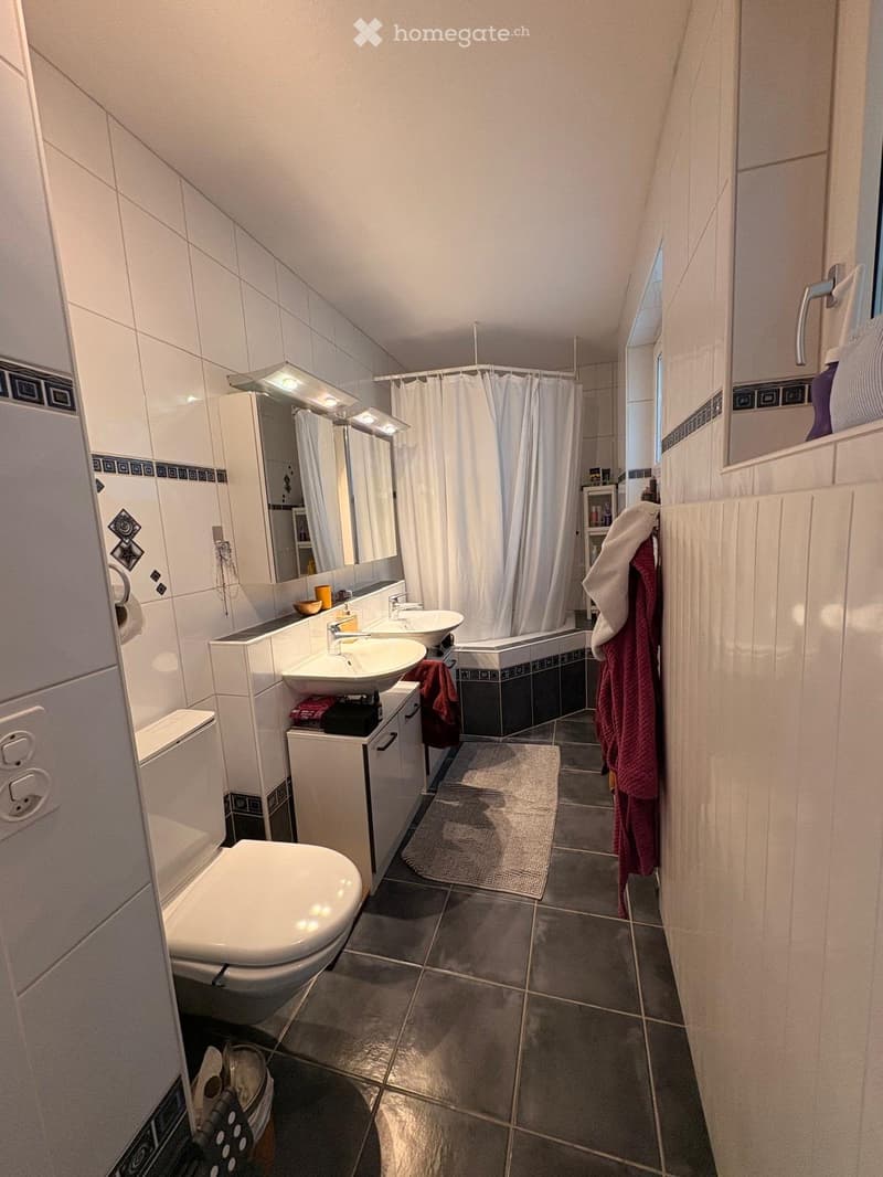 Modern bathroom with white tiles, a toilet, two sinks, a bathtub with a curtain, a mirror, and a towel rack with towels.