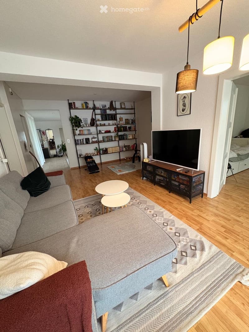 Modern living room with a grey sofa, ottoman, coffee table, small round table, television, bookshelf, and hanging pendant lights.