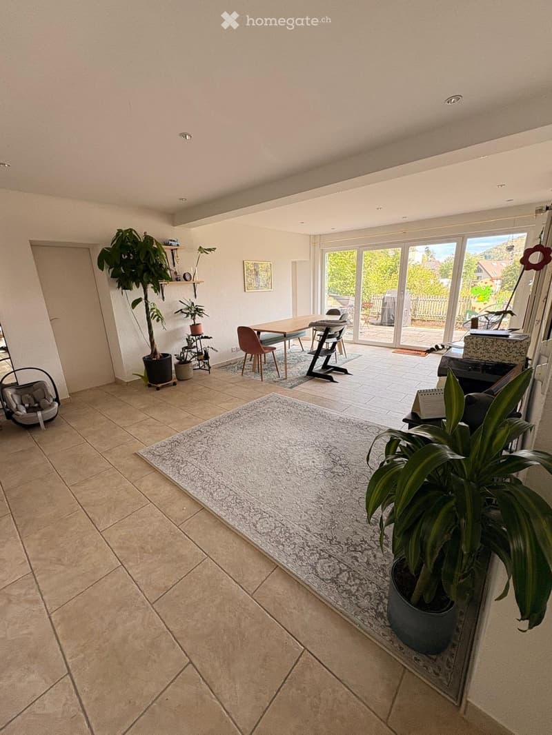 Bright living room with tiled floor, large potted plants, a sofa, a dining table with chairs, and a baby seat near the entrance.