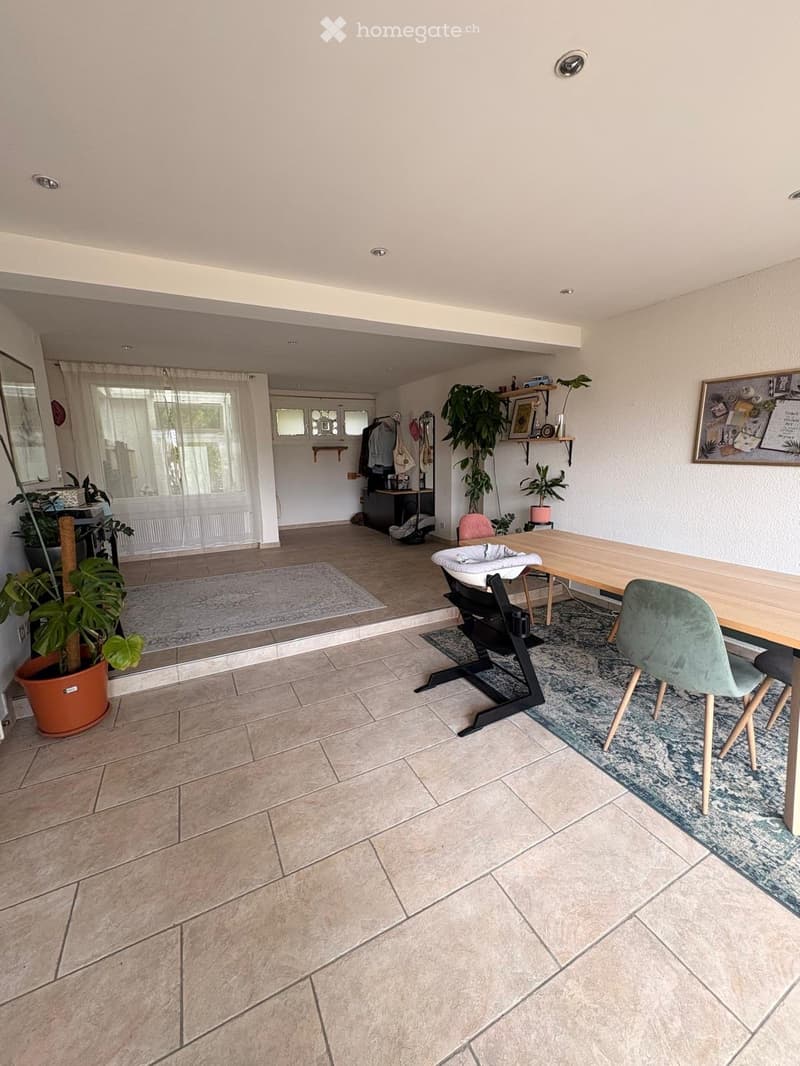 Bright, open living space with tiled flooring, large windows, and modern furniture including a dining table, chairs, baby chair, and decorative plants.
