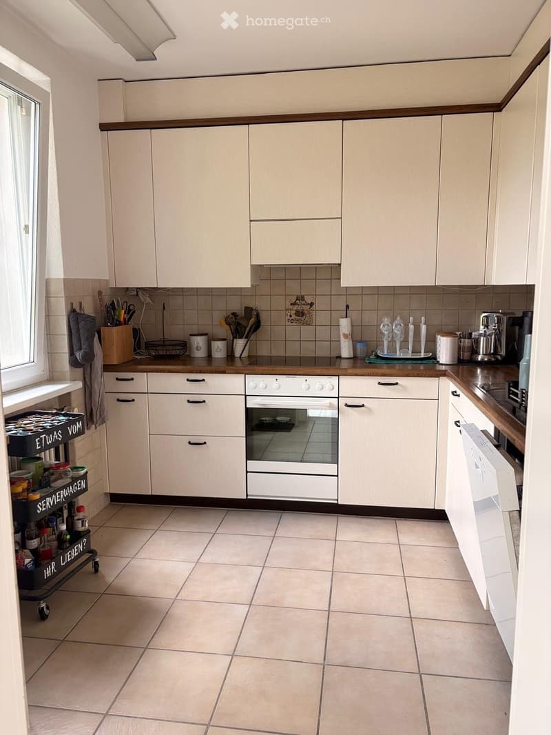 Modern kitchen with white cabinets, a central island, and various appliances including an oven and dishwasher. The countertop features a variety of kitchen items such as cups, bottles, and a kitchen cart with additional supplies.