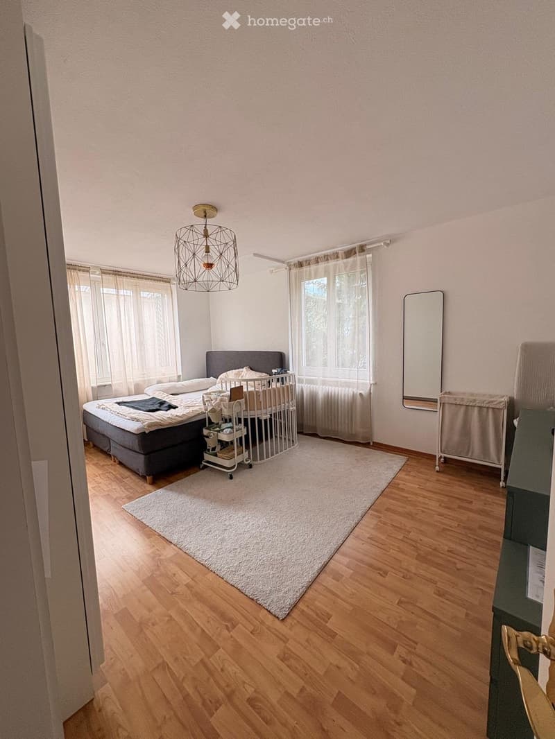 Bright bedroom with wooden flooring, large windows with sheer curtains, a double bed with pillows and blankets, a baby crib, a small cart with baby items, a large white rug, and a wall-mounted mirror.
