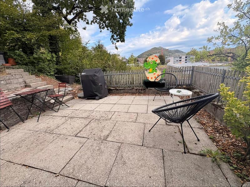 Outdoor patio with stone tiles, a small table, chairs, an umbrella, and a covered grill. Surrounded by greenery and a wooden fence, with a scenic view of hills in the background.