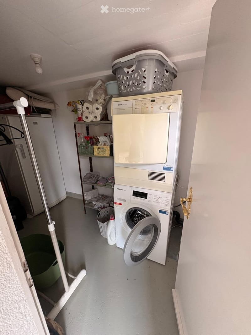 A small utility room with a stacked washing machine and dishwasher, shelves holding cleaning supplies, laundry baskets, and a broom and mop leaning against the wall.