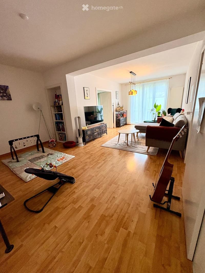 Spacious living room with wooden flooring, large windows, a sofa, coffee table, TV, bookshelf, and decorative items.