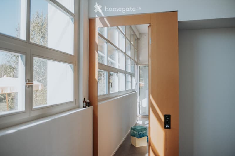 Bright hallway with large windows, light-colored walls, and a small decorative stool in the corner.