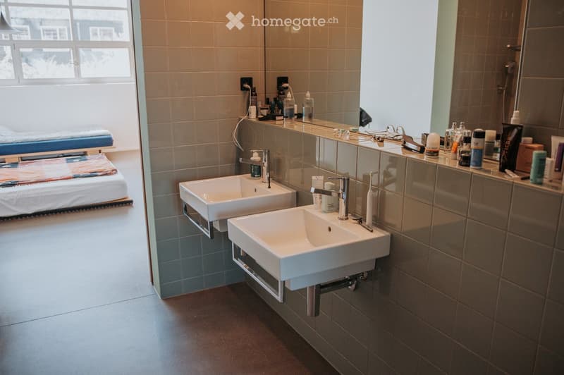 Modern bathroom with two white sinks, mirrors, and various toiletries on the counter. The room features tiled walls and a large window letting in natural light.