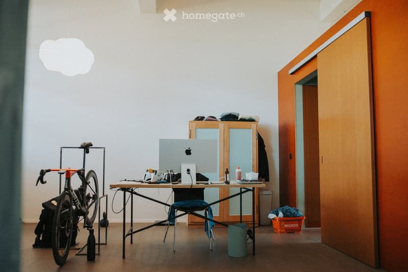 A modern, minimalist room with a wooden floor, white walls, and a large pendant light. A bicycle is mounted on a stand, a table holds a laptop and various items, and a cabinet with glass doors is visible.