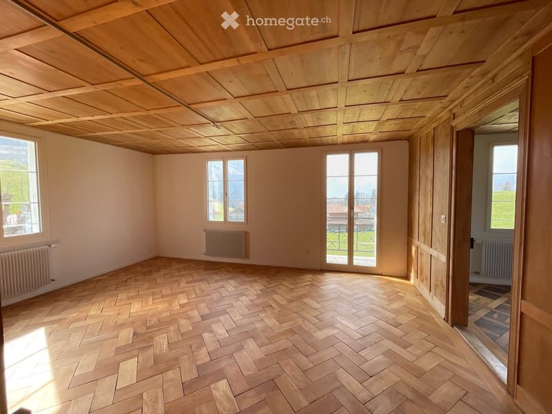 Empty room with wooden herringbone flooring, white walls, wooden ceiling, large windows, and radiators.